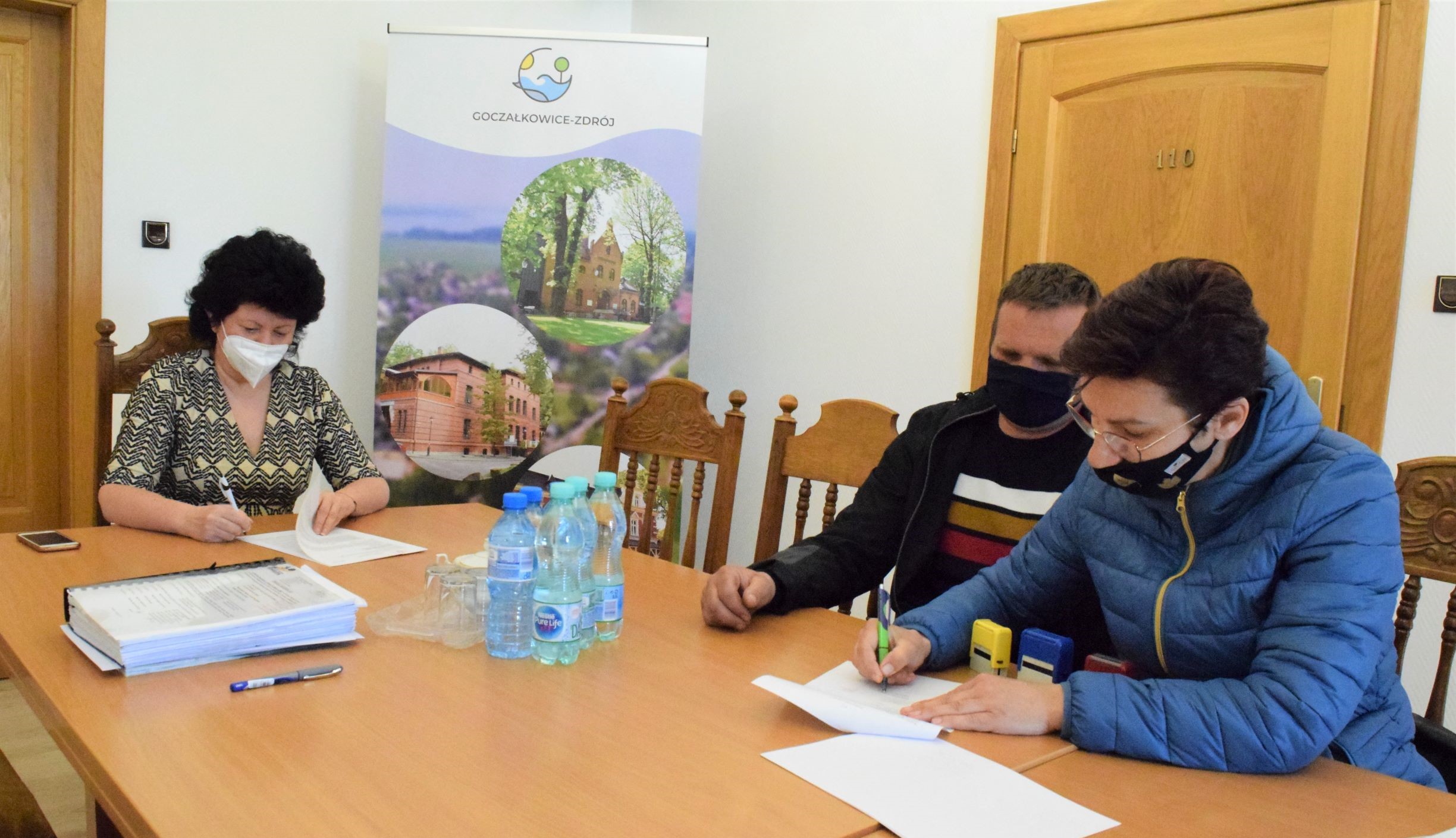 Zdjęcie przedstawia trzy osoby siedzące przy stole i podpisujące umowę